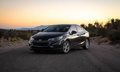 Black Chevrolet sedan parked on desert road at sunset with mountains and trees in background
